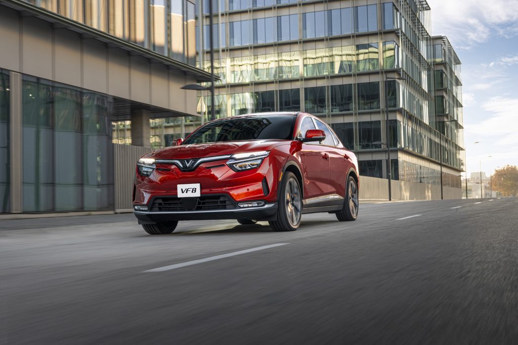 Red compact SUV (VF8) driving on a city street with modern glass-fronted office buildings in the background.